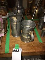Photo showing tall pewter mug with handle and attached lid next to a pewter mug with handle and ladle inside, placed on wooden surface with green tape markings.
