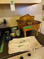 Wooden foot stool with floral seat and pair of Dutch wooden shoes placed on it, linens with crochet edges on counter, multiple ceramic bowls and cups visible