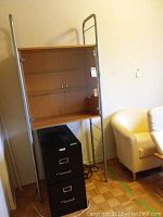Shelving unit consisting of wooden back panel with glass shelf and metal frame sides, located above the filing cabinet near an armchair.