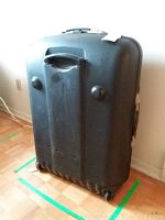 Full view of the black hard shell suitcase standing upright on four wheels, showing side handles and scuffs.