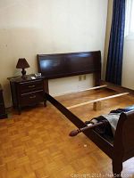 Dark wooden sleigh style bed frame partially assembled against wall with night table and lamp to the left
