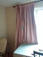 Pink/rose colored blackout curtains hanging with folds on a curtain rod, beside a window with surrounding wall and part of furniture visible.