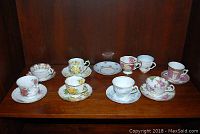 Eight different teacup and saucer sets displayed on wooden shelf showing assorted floral patterns and designs.