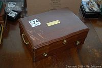 Closed wooden jewellery box with gold-tone handles and nameplate on the lid showing surface wear and scratches.