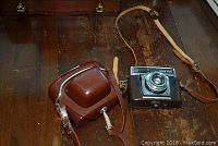 Photo showing Zeiss Ikon Contessamat camera next to its brown leather carrying case on a wooden surface.