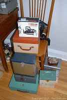 Stack of multiple metal and leather vintage camera/storage boxes of various colors and sizes on a wooden chair and floor.