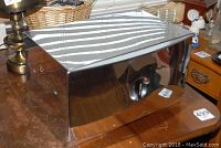 Front side of a polished chrome vintage bread box with an oval window and push-button mechanism.