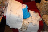 Various folded white and cream colored tablecloths and quilts, including embroidered and lace pieces, stacked on a red couch with a blue bag containing more linens.