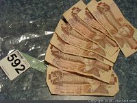 Seven 1986 Canadian two dollar bills displayed fanned out showing front side with Queen Elizabeth II, placed next to a clear plastic bag with a label