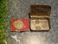 1922 USA one dollar coin in red plastic case alongside Evans Pastilles metal tin containing three 1943 steel pennies and a 1935 three pence coin on grey surface