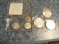 Six vintage Canadian coins displayed on countertop including three one dollar coins from 1963, 1964, and 1966; 1967 nickel; and two 1967 quarters. Coins show wear and are in plastic sleeves or clear display case.
