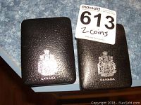 Two black hard display cases bearing the Canadian insignia, open to reveal a Canadian one dollar silver coin from 1971 and 1972. Cases are on a blue speckled surface.