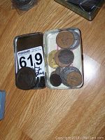 Photo showing a tin box and a small container with several coins of various sizes and colors including copper and silver tones on a wooden floor.