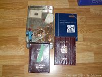 Coins and coin collection books on wooden floor, with loose pennies in plastic container and small figurine on metal tray.