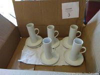 5 tall cylindrical porcelain cups and 6 round saucers placed inside a cardboard box on a cloth.