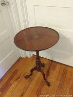 Full view of the vintage tilt-top round side table with a single pedestal base and ball and claw feet on a hardwood floor beside white doors.