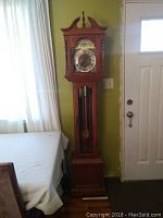 Full view of tall wooden grandfather clock standing next to door and near window with curtains.