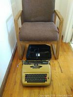 Brown fabric chair with wooden armrests and frame, hardwood floor background, and Sears Electric 1 beige electric typewriter on the floor with black plastic carrying case behind it.