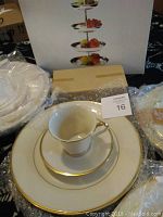 View of china set with stacked items including cup, saucer, dinner plate, some wrapped in plastic, and boxed 4-tier dessert pedestal in background