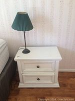White painted night stand with two drawers and black metal table lamp with pleated dark green shade, placed beside a bed, lit by natural light