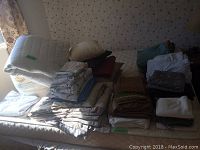 Photo of a variety of folded bed linens, blankets, pillows arranged on a bed, showing quantity and assortment