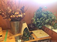 Photo shows dried flowers in large metal vase, a smaller beige vase, brown jug lamp, and faux ivy plant against an orange wall.