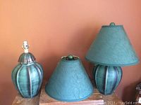 Two green table lamps on a wooden surface, one with a shade and one without the shade. Shades are green fabric. One shade shows a slight burn mark on top.