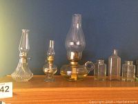 Nine glass pieces on wooden shelf including three oil lamps with glass chimneys and six small decorative glass bottles of various shapes and sizes.