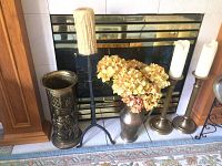 Photo of brass umbrella stand, iron candle stand with candle, brass candle holders with white candles, and faux floral arrangement in brass vase placed in front of fireplace.