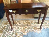 Front view of the wooden accent table showing three drawers and curved legs on a patterned rug.