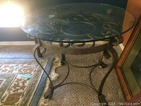 Side angle view showing the glass top and metal frame with scrollwork design on carpeted floor.