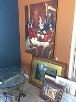 Photo showing large wall art with cello players and guitar, framed prints and art stacked beside it, glass side table partially visible.
