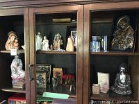 Photo showing dark wood display cabinet with glass doors housing various religious and Asian decorative items including statues, figurines, and artwork.