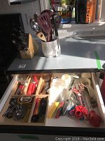 Top view of kitchen countertop showing wooden knife block with three Henckels knives, white vase holding kitchen utensils, and partially open drawer with kitchen tools.