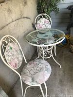 Glass top bistro table with white metal frame and two white metal chairs with floral cushions