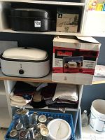 Photo showing a GE white oval food server with dial control, a blue basket full of glass canning jars with lids, some folded linens or towels, and a black speckled roaster pan on shelf above.