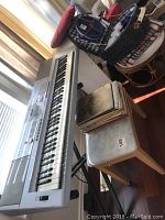 Full side view of Yamaha DGX203 keyboard on adjustable stand with matching silver bench and a stack of sheet music beside keyboard bench.