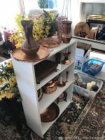 White shelving unit holding multiple copper items including bowls, plates, warmer, kettle, and vase with yellow flowers beside it.