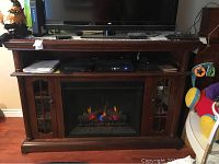 Front view of the media center with a large flat-screen TV on top, visible electronic devices on the shelf above the fireplace, and the fireplace lit with realistic flame effects.