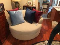 Front view of the large circular beige rotating upholstered chair with four pillows in blue and red shades on it.