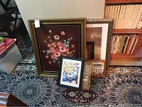 Shows multiple framed pictures leaning against wall, including large floral painting and smaller pieces, placed on patterned carpet