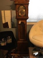 Full view of the tall grandfather clock next to a white chair showing its wooden case and overall size.