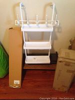 White metal hanging shelf rack with three shelves and hooks leaning against the wall with cardboard boxes on both sides.