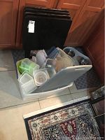 Four black folding TV trays standing against wooden cabinets, with a large plastic bin filled with assorted plastic containers in front of them, placed on a tiled floor next to area rugs and pet food bowls.