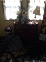 Chest of drawers with lamp on top, artificial flower arrangement, umbrellas, and rugs on floor in front of chest