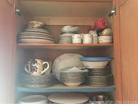 Photo showing stacked dinnerware inside a cabinet, including plates, bowls, cups, and saucers from both Noritake and Bellavino sets.