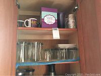 Inside wooden cabinet shelf showing 8 tall clear glass tumblers, 8 short clear glass tumblers, assorted ceramic coffee cups in various colors and designs.