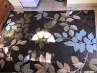 Brown area rug with floral leaf patterns in light blue, beige, and olive green on a dark brown background, shown on tile floor near wooden furniture.