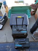 Photo showing two soft black suitcases, one with a securing strap, and several plastic storage containers and photo boxes of different sizes and colors stacked on the ground.