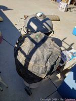 Black charcoal BBQ grill covered with a worn black cover, lid partially visible behind cover, grill on wheels outside on concrete surface.